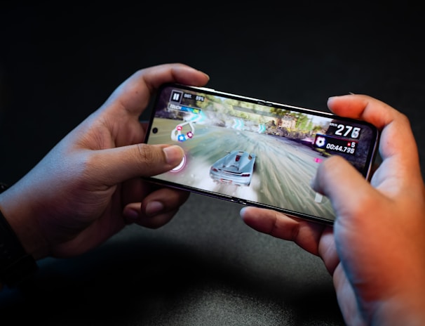 Hands holding a smartphone with a racing game on the screen, featuring a car speeding along a track with visible game controls and speed indicators.